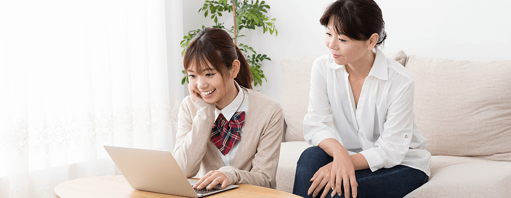 ソファに座りながらノートパソコンを見る女子学生と隣にいる母親