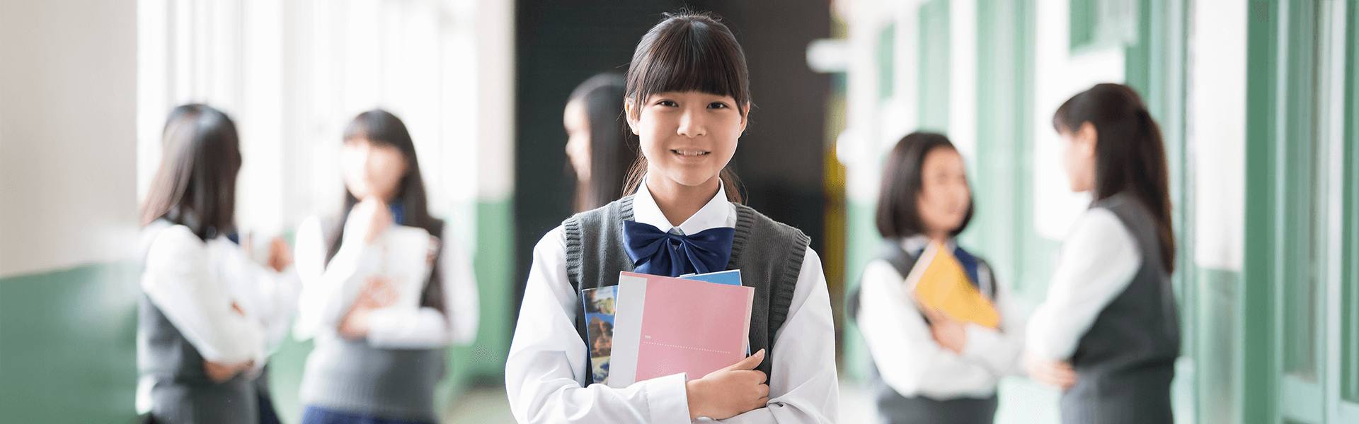 廊下で本を抱えて立つ女子中学生と周囲で会話する生徒たち