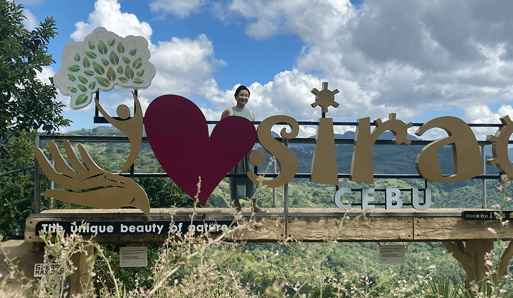 笑顔の女性が「i ♥ sirao」のサインの後ろで自然の景色を背景に立っている