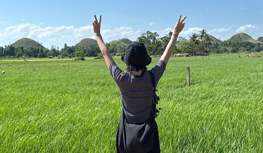 ストライプシャツを着た人物が緑の田んぼで両腕を上げてピースサインをしている屋外の自然景色