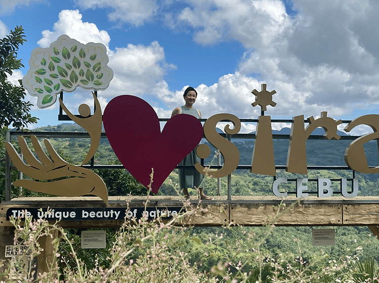 笑顔の女性が「i ♥ sirao」のサインの後ろで自然の景色を背景に立っている