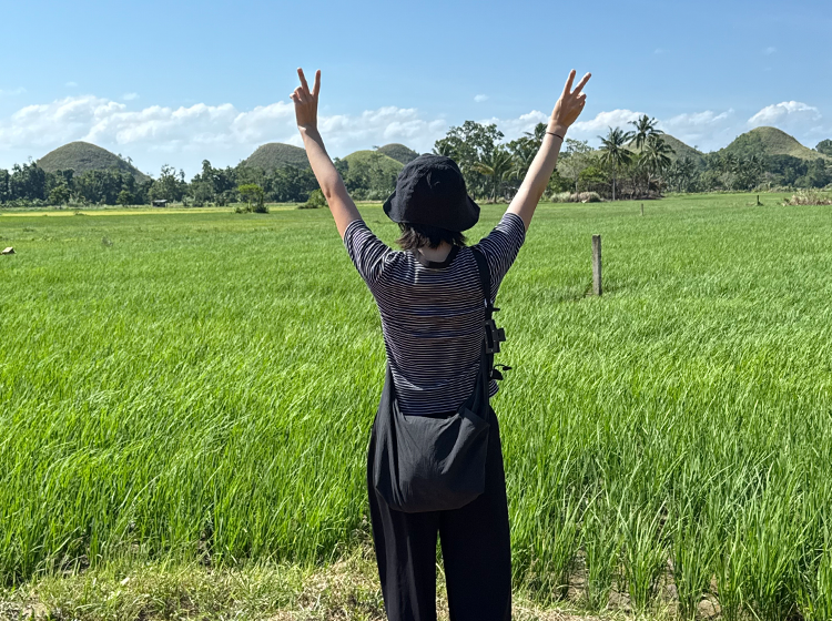 ストライプシャツを着た人物が緑の田んぼで両腕を上げてピースサインをしている屋外の自然景色